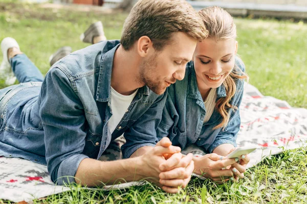 Sorrindo jovem casal deitado na grama no parque e usando smartphone — Fotografia de Stock