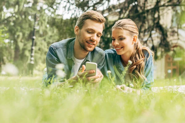 Happy young couple lying on grass at park and using smartphone — Stock Photo