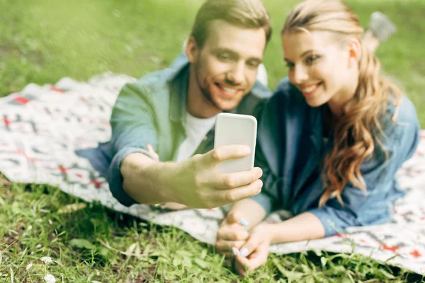 Beautiful young couple lying on grass at park and taking selfie — Stock Photo