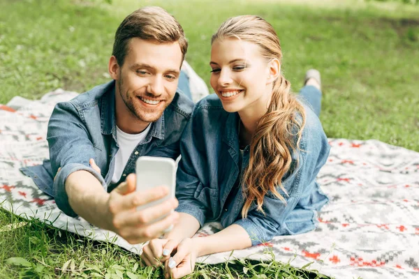 Smiling young couple lying on grass at park and taking selfie — Stock Photo