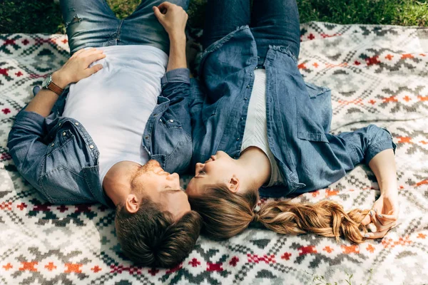 Высокий угол обзора улыбающейся молодой пары, лежащей на узорчатой ткани на траве — стоковое фото
