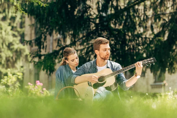 Attraente giovane uomo che suona la chitarra per la sua ragazza durante il picnic al parco — Foto stock