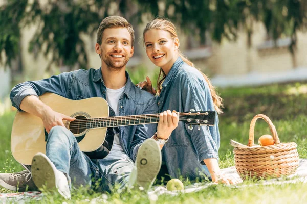 Felice giovane uomo che suona la chitarra per la sua ragazza sorridente durante il picnic al parco e guardando la fotocamera — Foto stock