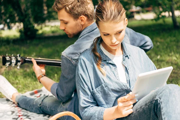 Beau jeune homme jouant de la guitare pour sa petite amie alors qu'elle utilise une tablette sur l'herbe au parc — Photo de stock