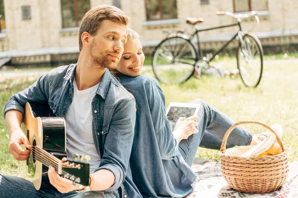 Bel giovane uomo che suona la chitarra per la sua ragazza sorridente mentre fa un picnic al parco — Foto stock