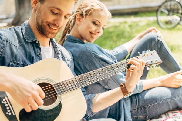 Schöner junger Mann spielt Gitarre für seine lächelnde Freundin, während er sich im Park entspannt — Stockfoto