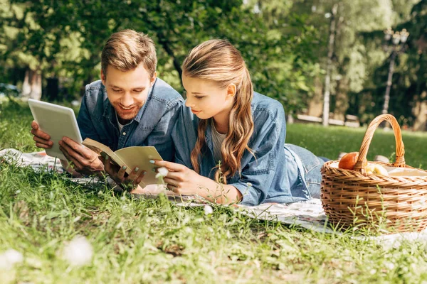 Feliz joven pareja acostada en la hierba en el parque con la tableta y el libro - foto de stock