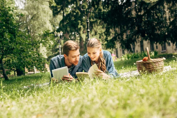 Schönes junges Paar liegt mit Tablet und Buch im Park auf Gras — Stockfoto