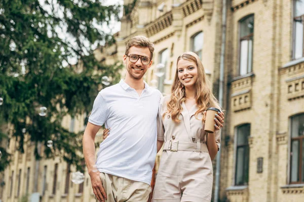 Sonriente joven pareja en elegante ropa mirando a la cámara en frente de viejo edificio - foto de stock