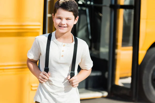 Pequeño alumno sonriente con mochila de pie frente al autobús escolar - foto de stock