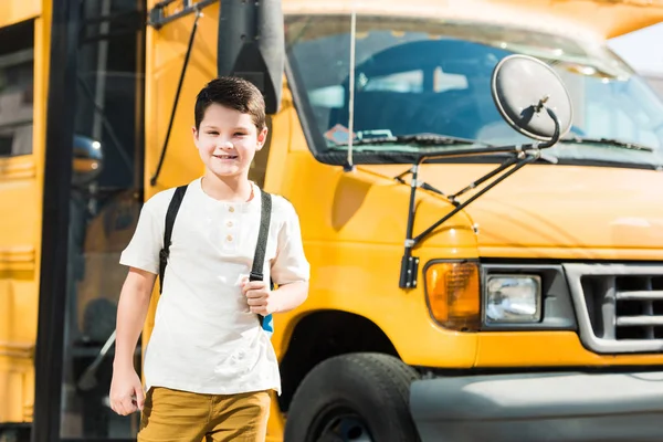 Heureux petit élève avec sac à dos debout devant le bus scolaire — Photo de stock