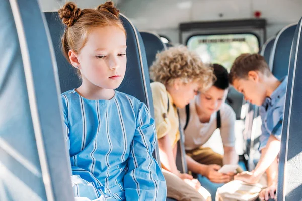 Пригнічена маленька школярка сидить одна в шкільному автобусі, в той час як її однокласники використовують смартфон разом на фоні — стокове фото