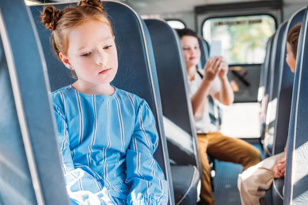 Traurige kleine Schülerin, die allein im Schulbus sitzt, während ihre Klassenkameraden im Hintergrund plaudern — Stockfoto