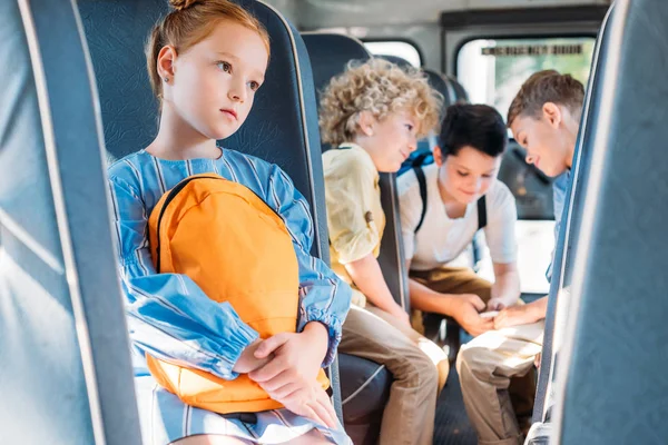 Petite écolière déprimée assise seule dans le bus scolaire pendant que ses camarades de classe s'amusent ensemble sur fond — Photo de stock