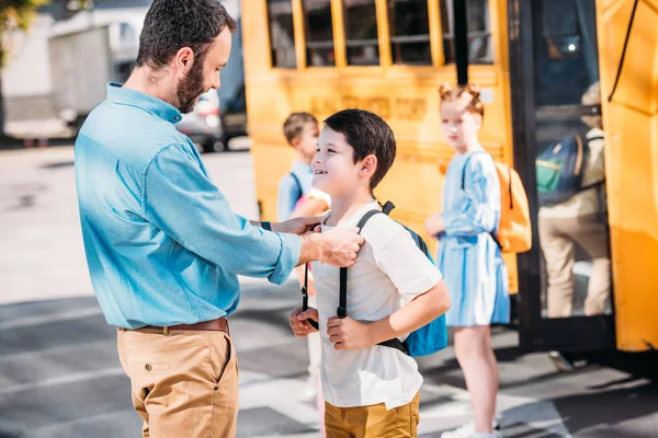 Padre guapo hablando con su hijo antes de la escuela en frente del autobús escolar con sus compañeros de clase - foto de stock