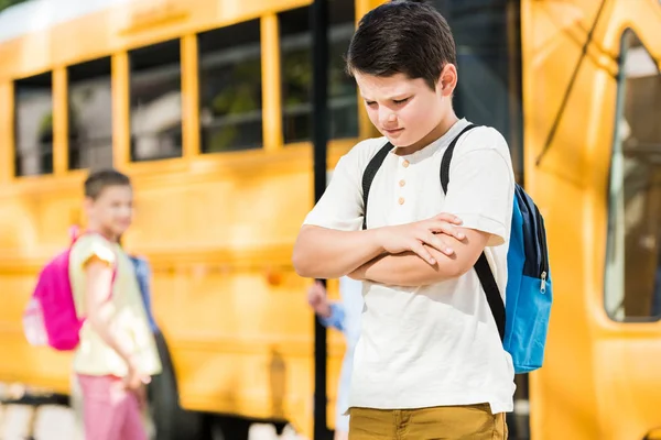 Depressed little schoolboy with crossed arms standing in front of school bus — Stock Photo
