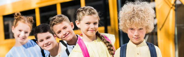 Panoramaaufnahme einer Gruppe entzückender Schüler, die in die Kamera schauen, während sie vor dem Schulbus in der Reihe stehen — Stockfoto