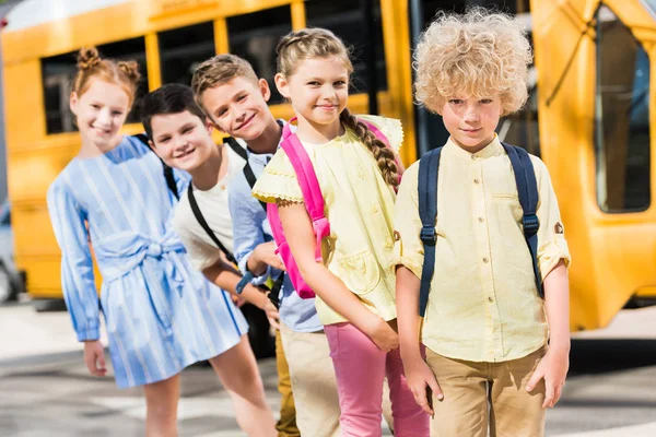 Gruppe entzückender Schüler blickt in die Kamera, während sie in der Reihe vor dem Schulbus steht — Stockfoto