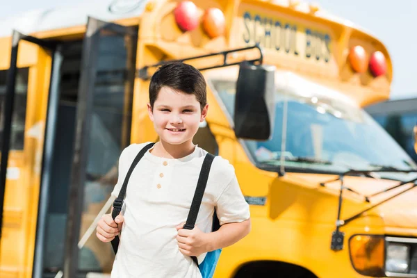 Pequeño colegial sonriente con mochila de pie frente al autobús escolar - foto de stock