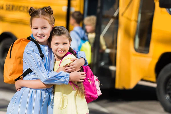 Beautiful happy schoolgirls looking at camera and embracing in front of school bus — Stock Photo