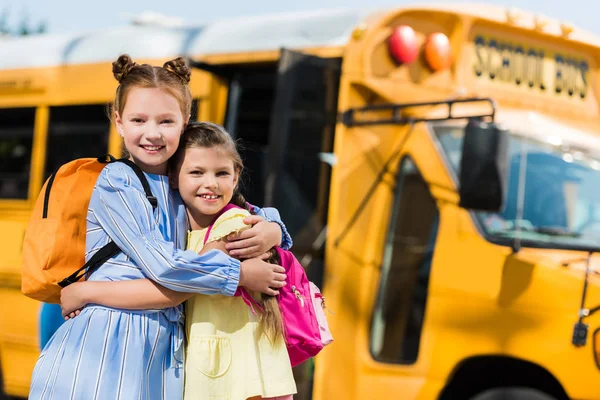 Счастливые маленькие школьницы обнимаются перед школьным автобусом и смотрят в камеру — стоковое фото