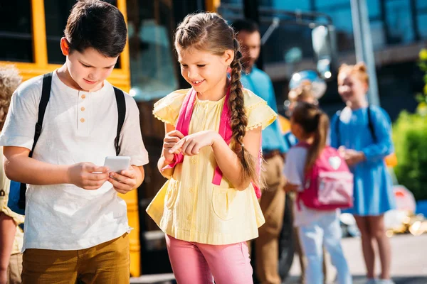 Happy schoolgirl and schoolboy using smartphone together in front of school bus with classmates — Stock Photo