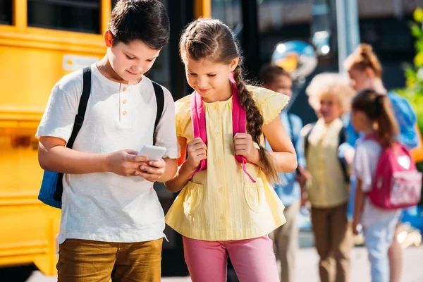 Schoolgirl and schoolboy using smartphone together in front of school bus with classmates — Stock Photo