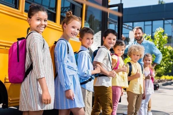 Coda di allievi felici in piedi vicino a scuolabus con insegnante — Foto stock