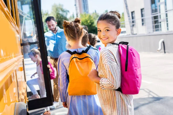 Pequena estudante entrando ônibus escolar com colegas de classe, enquanto professor de pé perto da porta — Fotografia de Stock