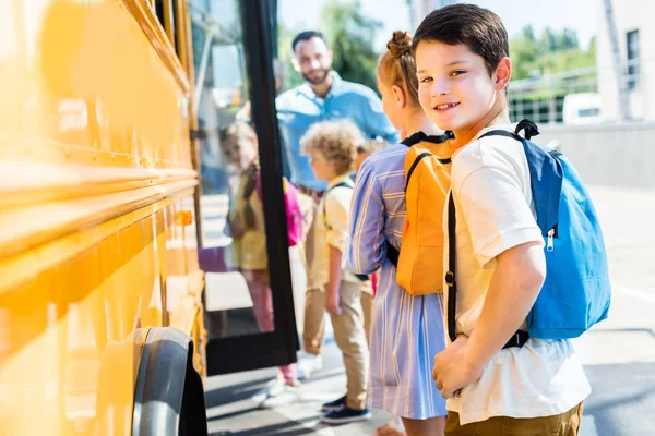 Маленький школьник входит в школьный автобус с одноклассниками, а учитель стоит рядом с дверью — стоковое фото