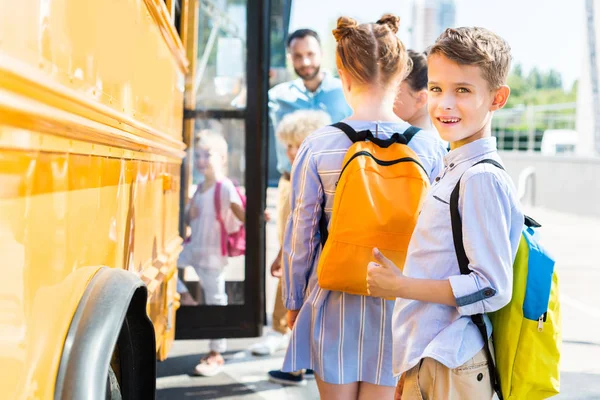 Усміхнений маленький школяр, що входить в шкільний автобус з однокласниками, в той час як вчитель стоїть біля дверей — стокове фото