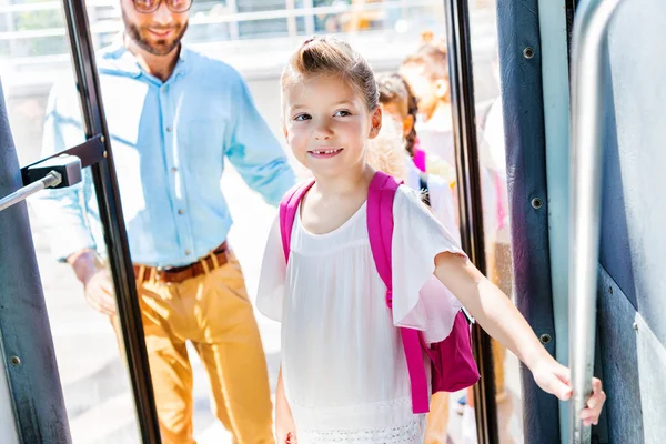 Красива маленька школярка, що входить в шкільний автобус з вчителем і однокласниками на фоні — стокове фото