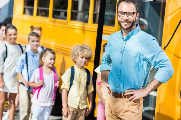 Bonito masculino professor olhando para câmera enquanto alunos entrando escola ônibus borrado no fundo — Fotografia de Stock