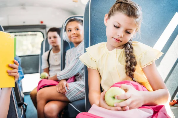 Beautiful little schoolgirl with backpack and apple riding on school bus with blurred classmates on background — Stock Photo