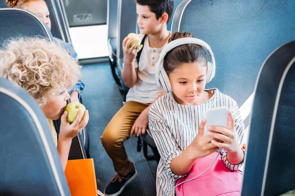 Adorável colegial usando smartphone e ouvir música com fones de ouvido enquanto andava de ônibus escolar com colegas de classe — Fotografia de Stock