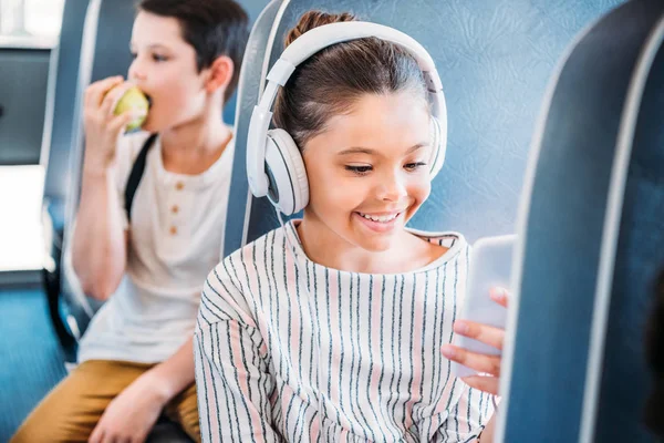 Scolaretta felice usando smartphone e ascoltando musica con le cuffie mentre cavalca sullo scuolabus — Foto stock