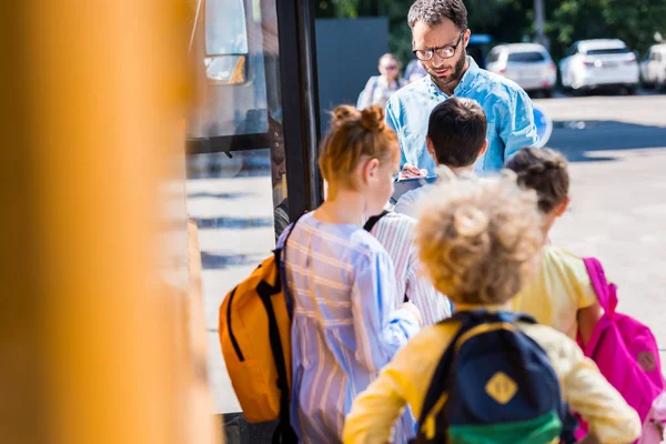 Vue arrière des élèves entrant dans l'autobus scolaire pendant que l'enseignant écrit dans le presse-papiers — Photo de stock