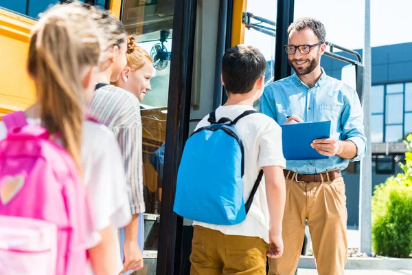 Pupils entering school bus while teacher writing in clipboard — Stock Photo