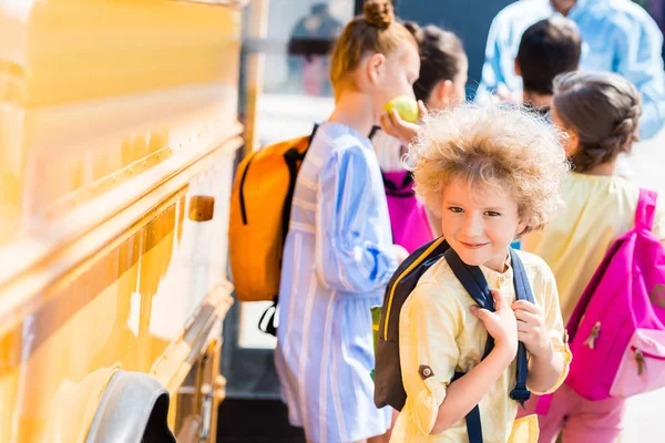 Щасливий кучерявий школяр зі своїми однокласниками, що стоять біля шкільного автобуса — стокове фото
