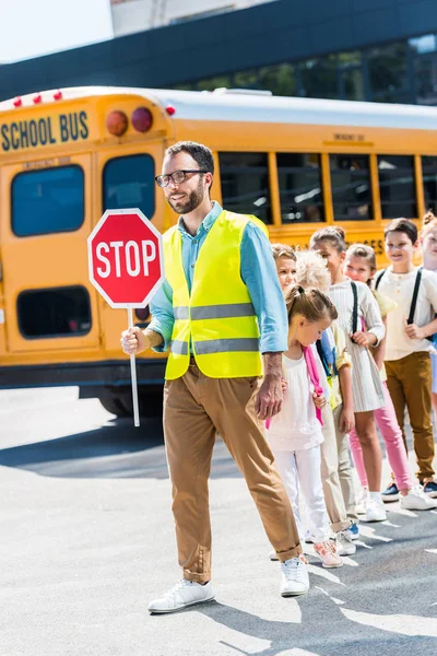 Guardia de tránsito cruzando la calle con alumnos felices delante del autobús escolar - foto de stock