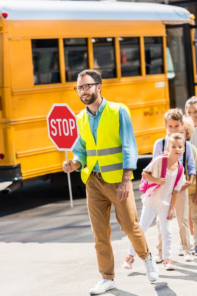 Trânsito feliz guarda cruzando estrada com alunos em frente ao ônibus escolar — Fotografia de Stock