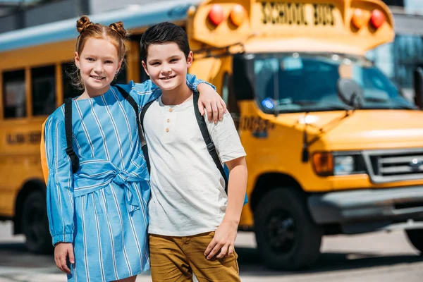 Счастливые маленькие ученые обнимаются и смотрят в камеру перед школьным автобусом — стоковое фото