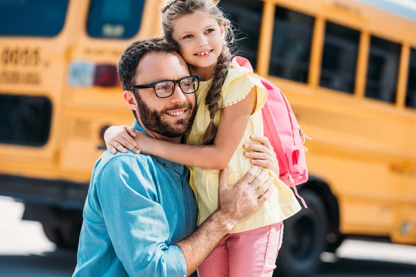 Красивый отец обнимает свою маленькую дочь перед школьным автобусом — стоковое фото