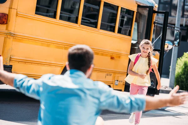 Vista trasera del padre con los brazos abiertos esperando a la pequeña y feliz hija mientras corre desde el autobús escolar - foto de stock