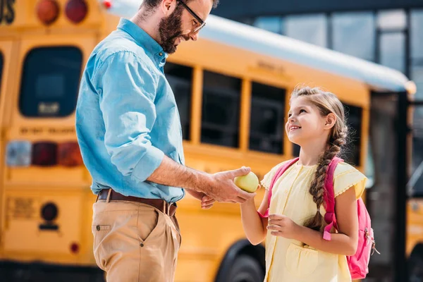 Père heureux donnant pomme verte fraîche à la fille devant le bus scolaire — Photo de stock