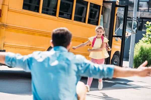 Padre con los brazos abiertos esperando a su hija mientras ella huye del autobús escolar - foto de stock