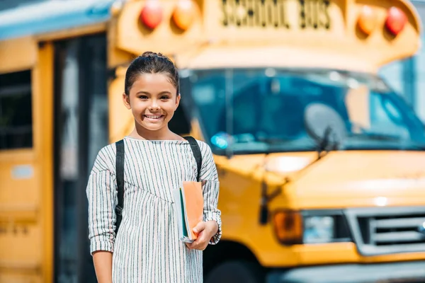 Adorable little schoolgirl with notebooks looking at camera in front of school bus — Stock Photo