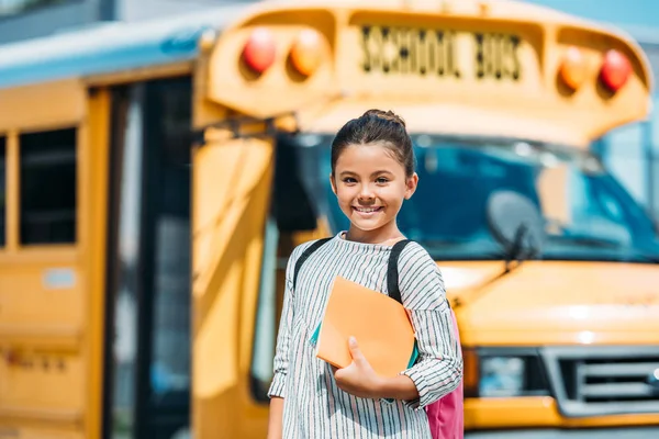 Feliz colegiala con cuadernos mirando a la cámara delante del autobús escolar - foto de stock