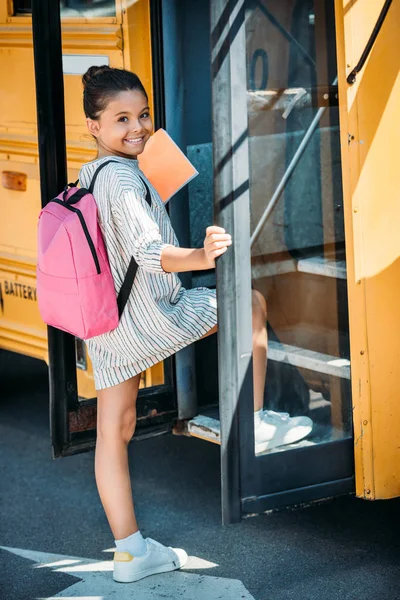 Adorable pequeña colegiala entrar en autobús escolar y mirando a la cámara - foto de stock