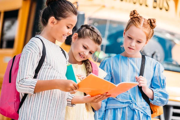 Grupo de colegialas mirando el cuaderno delante del autobús escolar - foto de stock